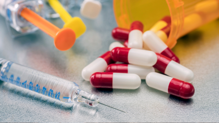 A syringe with a pill bottle and pill capsules in the background